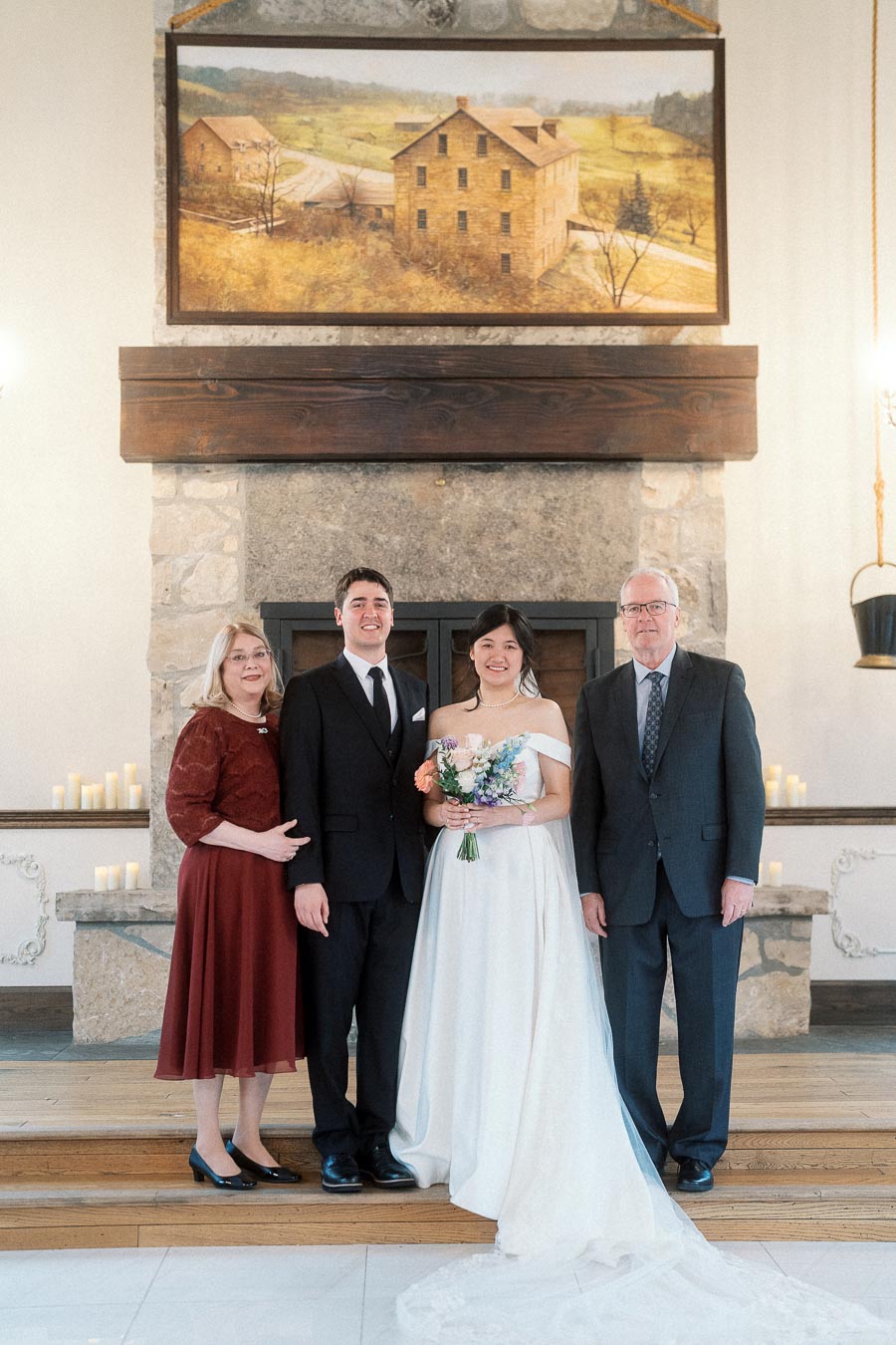 Bride and groom posing with two family members in front of a stone fireplace.