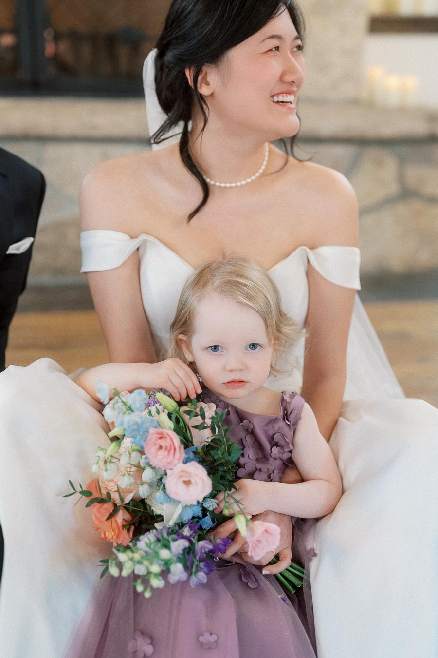 Woman in an off-the-shoulder white gown sitting with a child holding a colorful bouquet