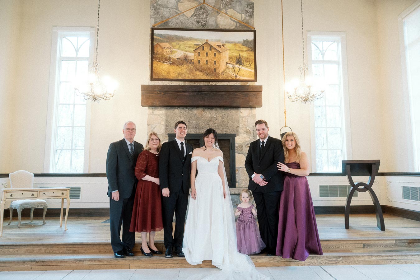 Formal group portrait with a bride, groom, family members, and child indoors by a fireplace