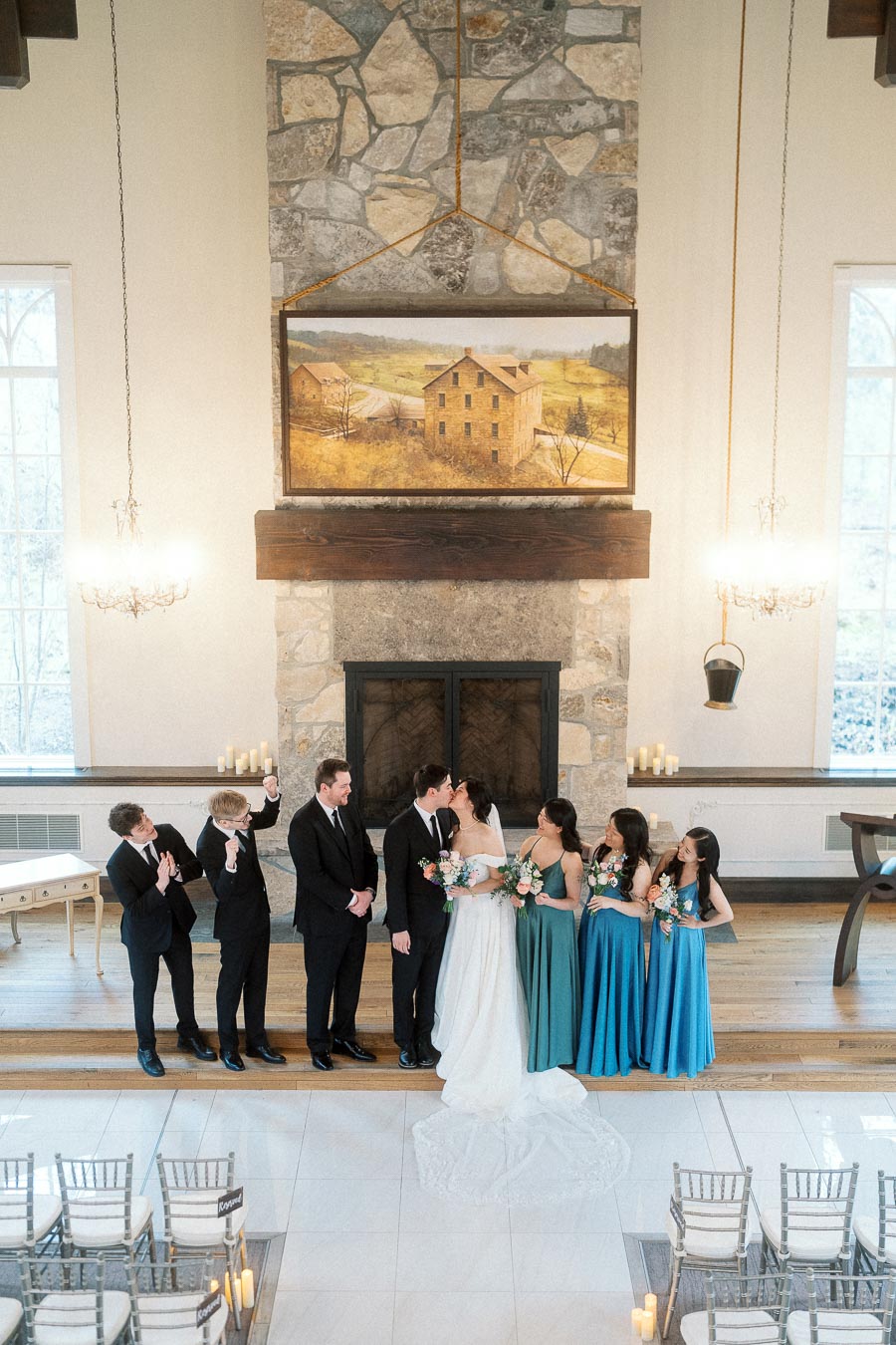 Wedding party standing indoors by a stone fireplace as the couple kisses