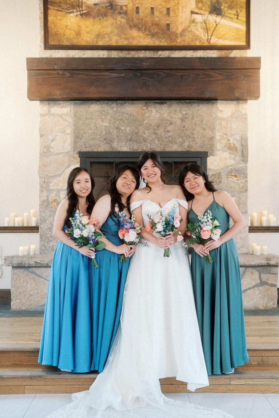 Bride posing with three bridesmaids holding bouquets in front of a stone fireplace