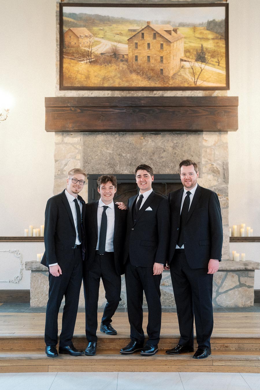 Four men in black suits standing together in front of a stone fireplace