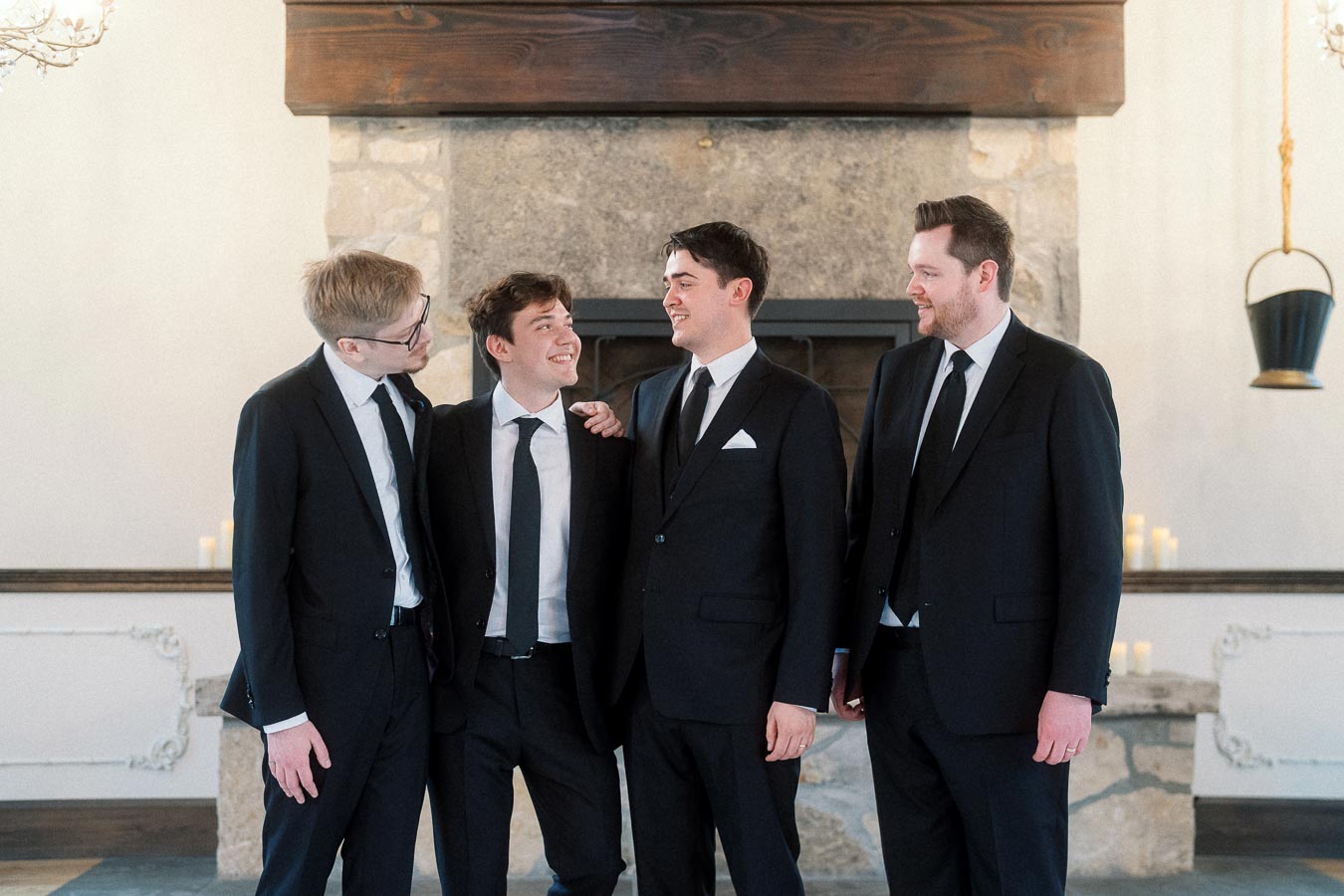 Four men in black suits smiling together in front of a stone fireplace