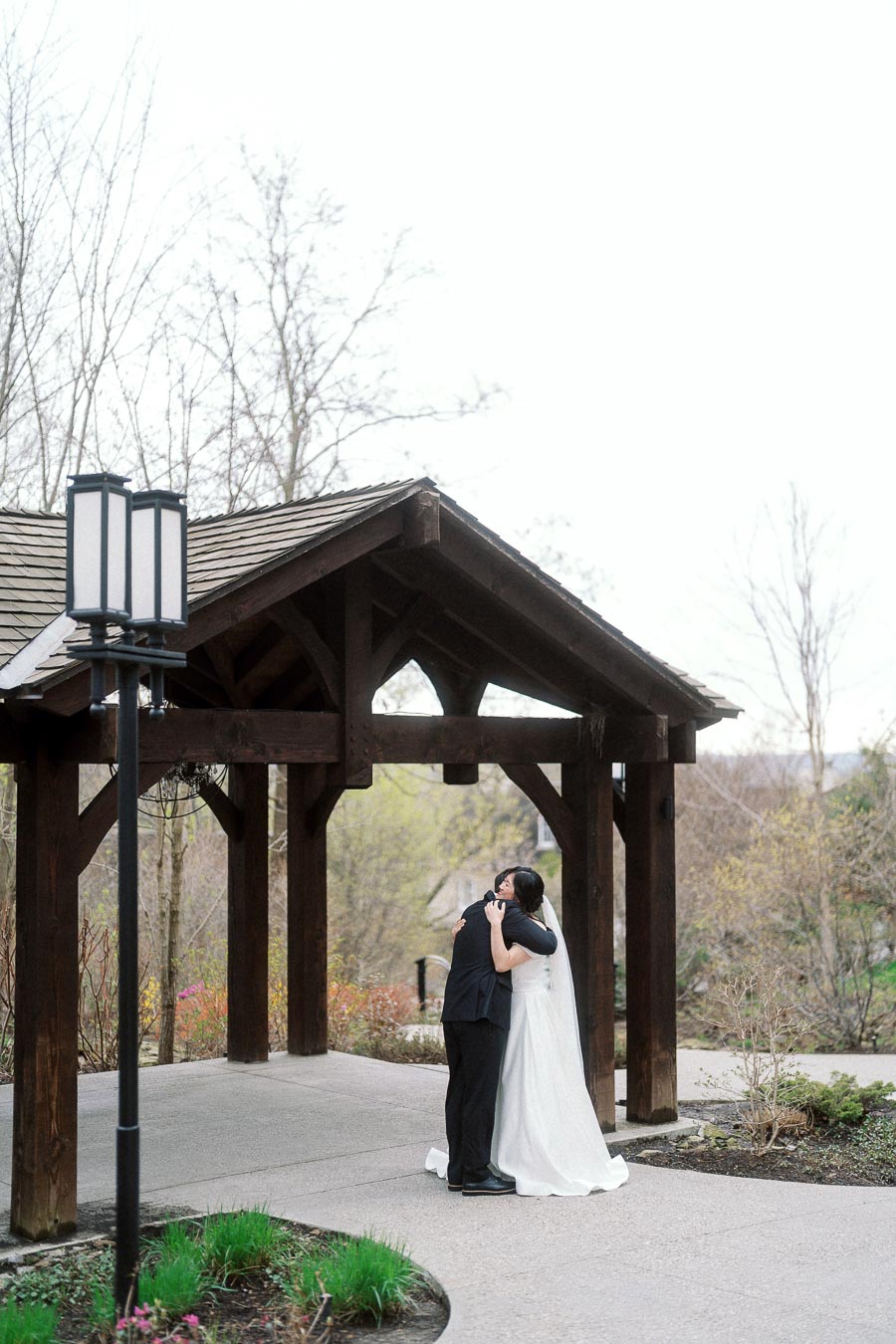 Couple in formal attire embracing beneath a wooden garden pavilion.