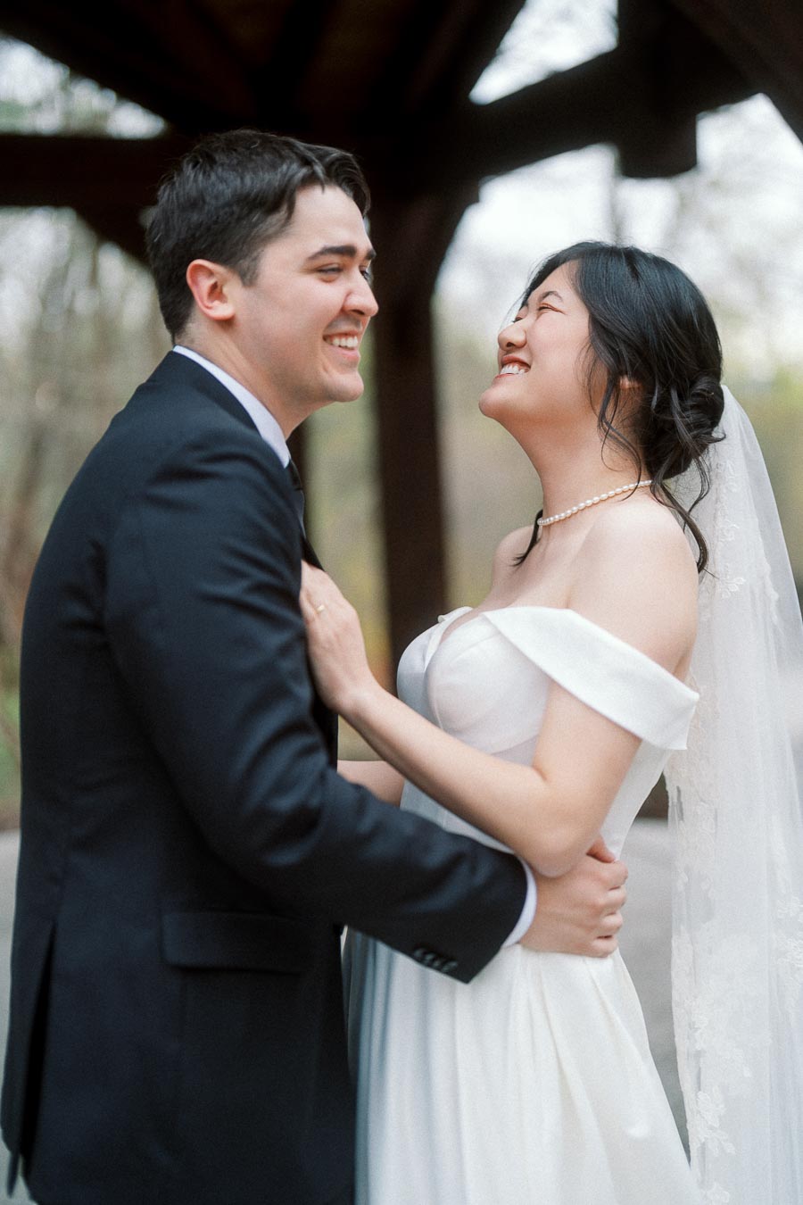 Couple in formal wedding attire laughing together under a wooden pavilion