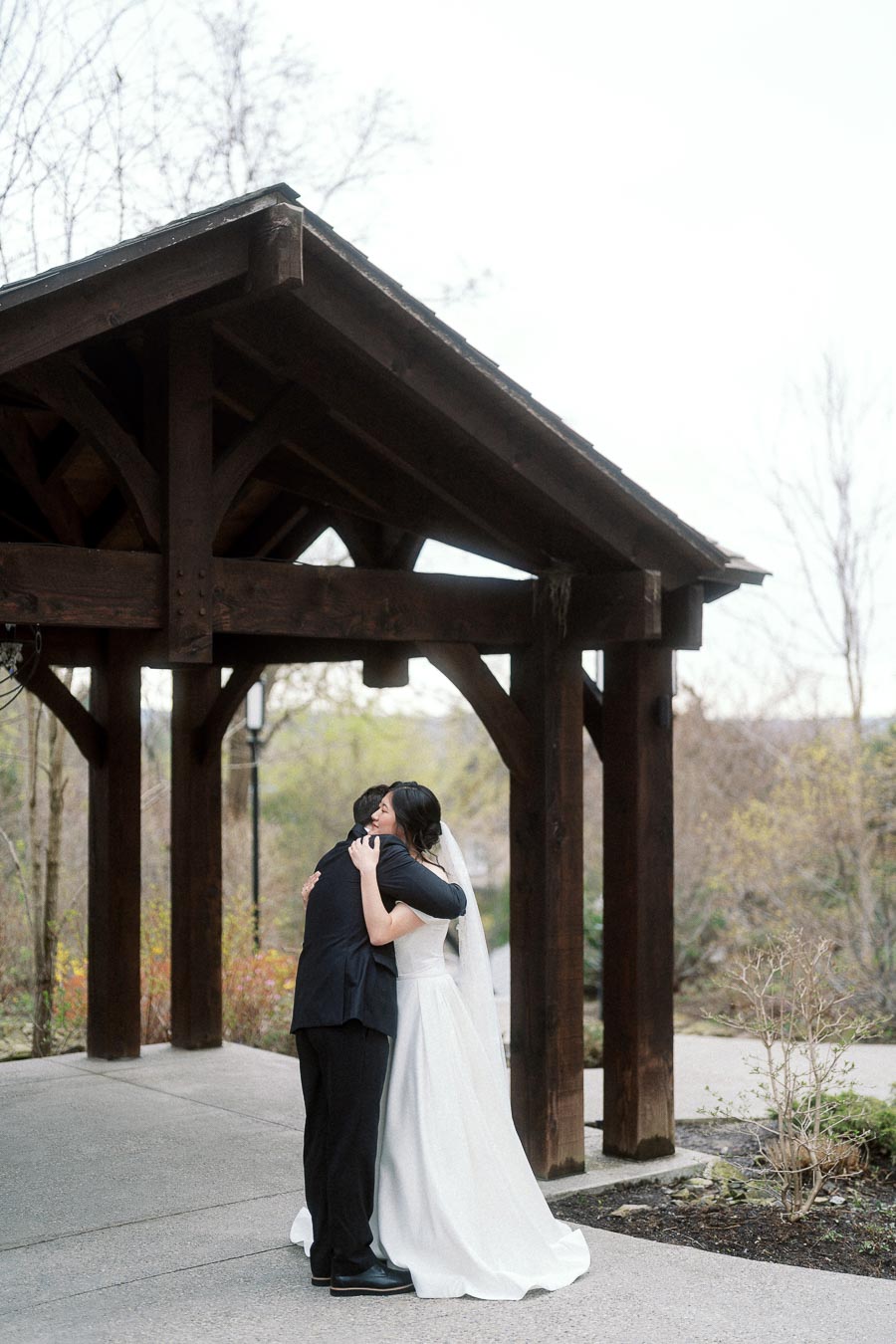 Couple in formal wedding attire embracing beneath a wooden pavilion outdoors