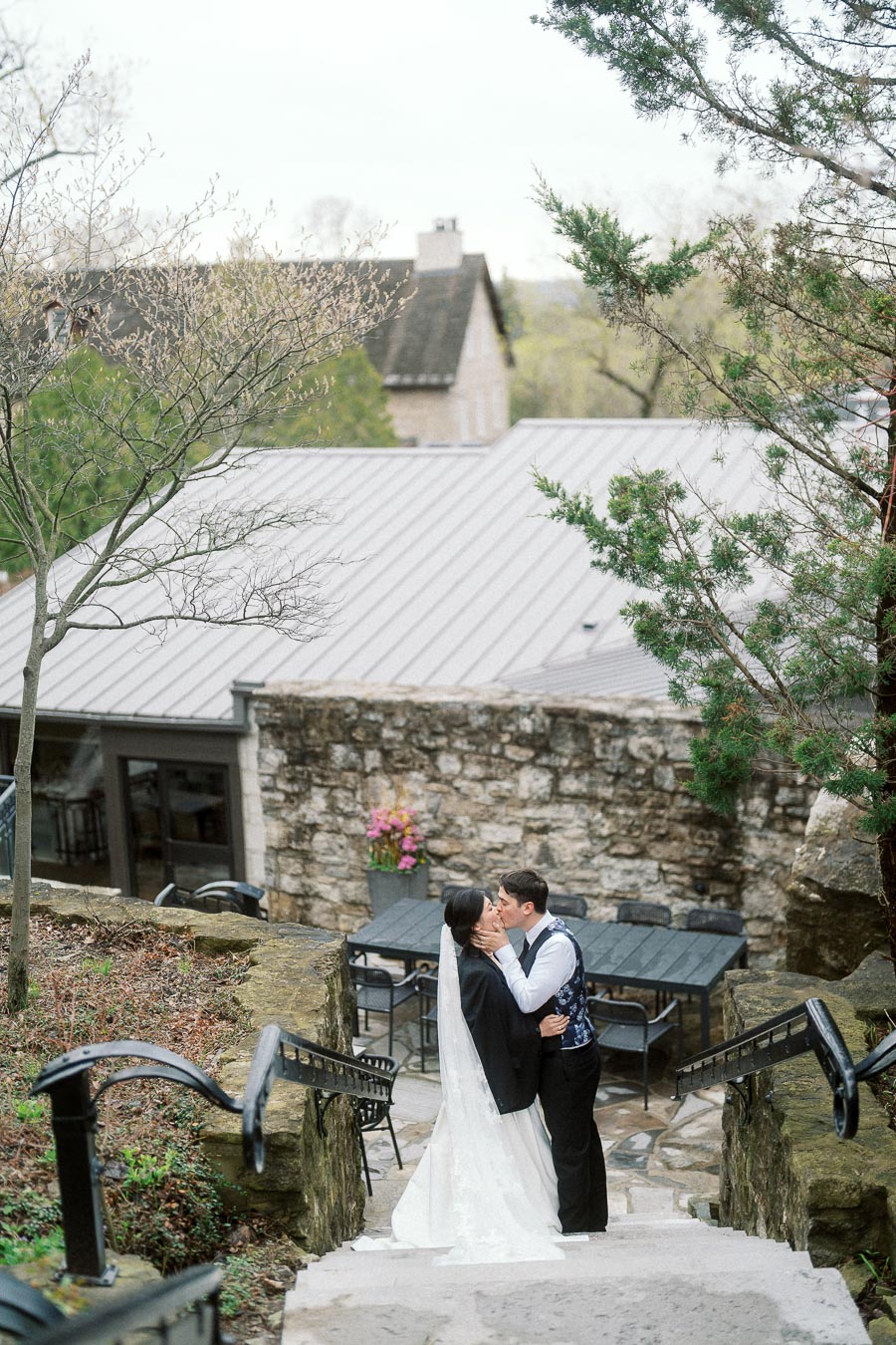 Couple in formal wedding attire kissing on stone steps near a rustic building