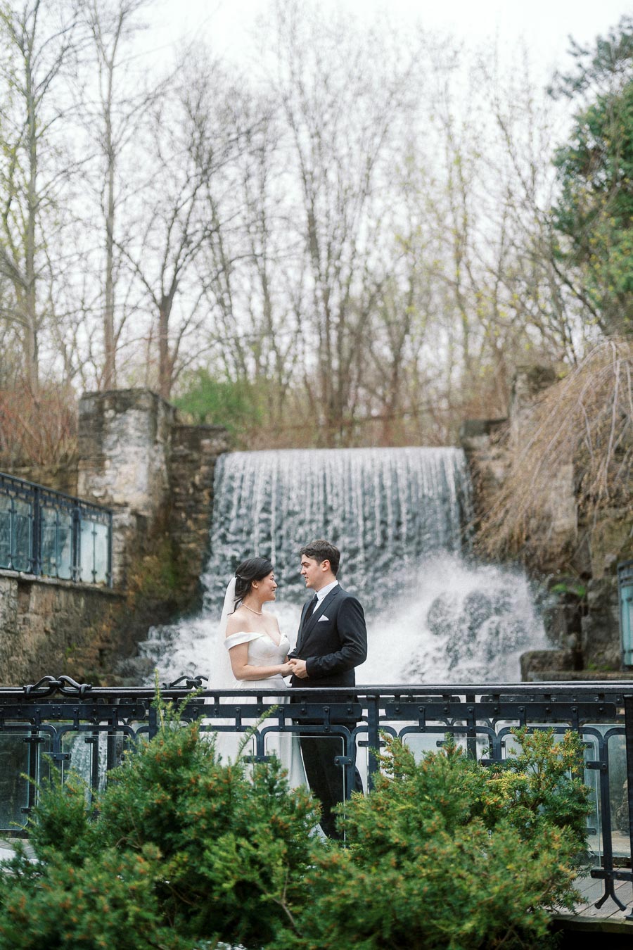 Couple in formal attire holding hands on a bridge in front of a waterfall