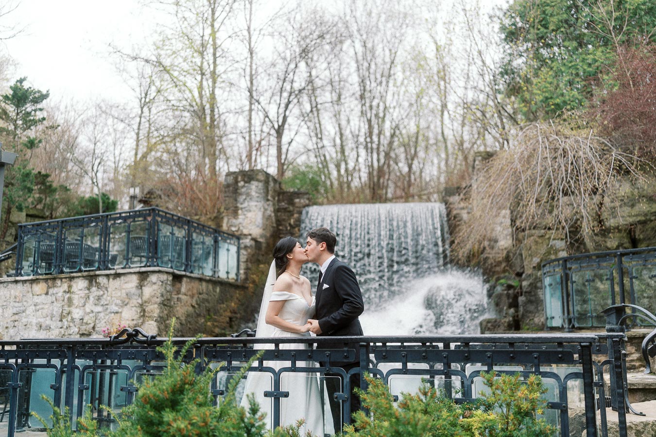 Couple in formal attire kissing beside a waterfall and railing