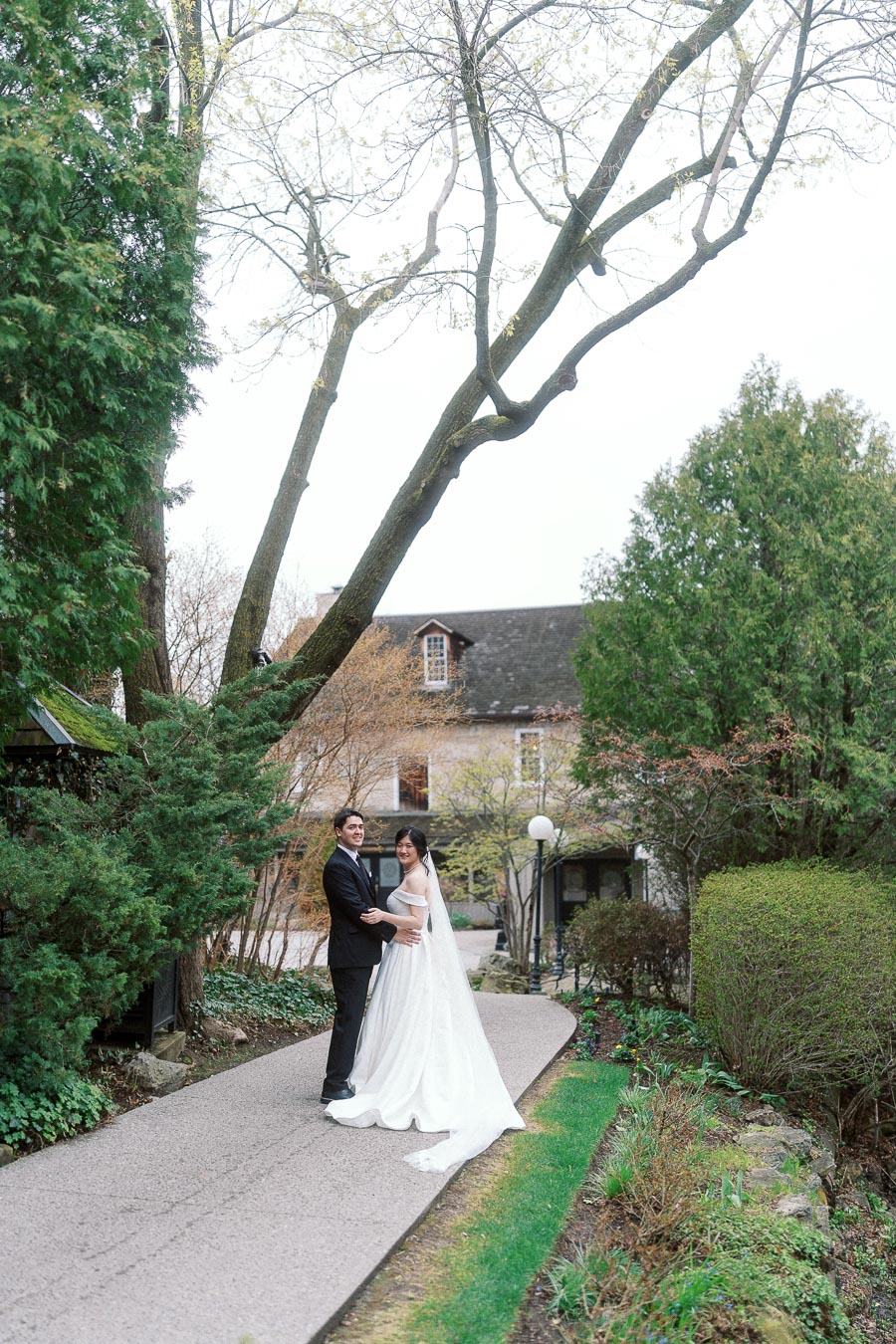 Couple in formal attire posing on a garden path beneath tall trees