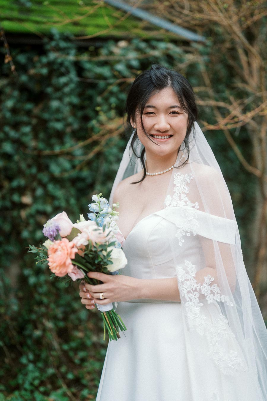 Smiling bride in a white dress and veil holding a pastel bouquet outdoors