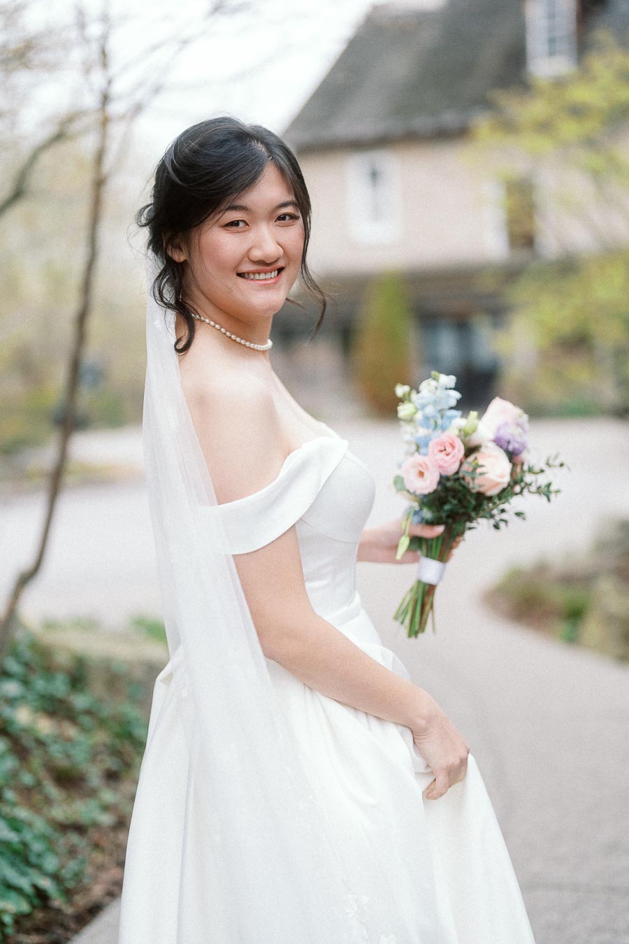Smiling woman in a white dress and veil holding a pastel bouquet outdoors
