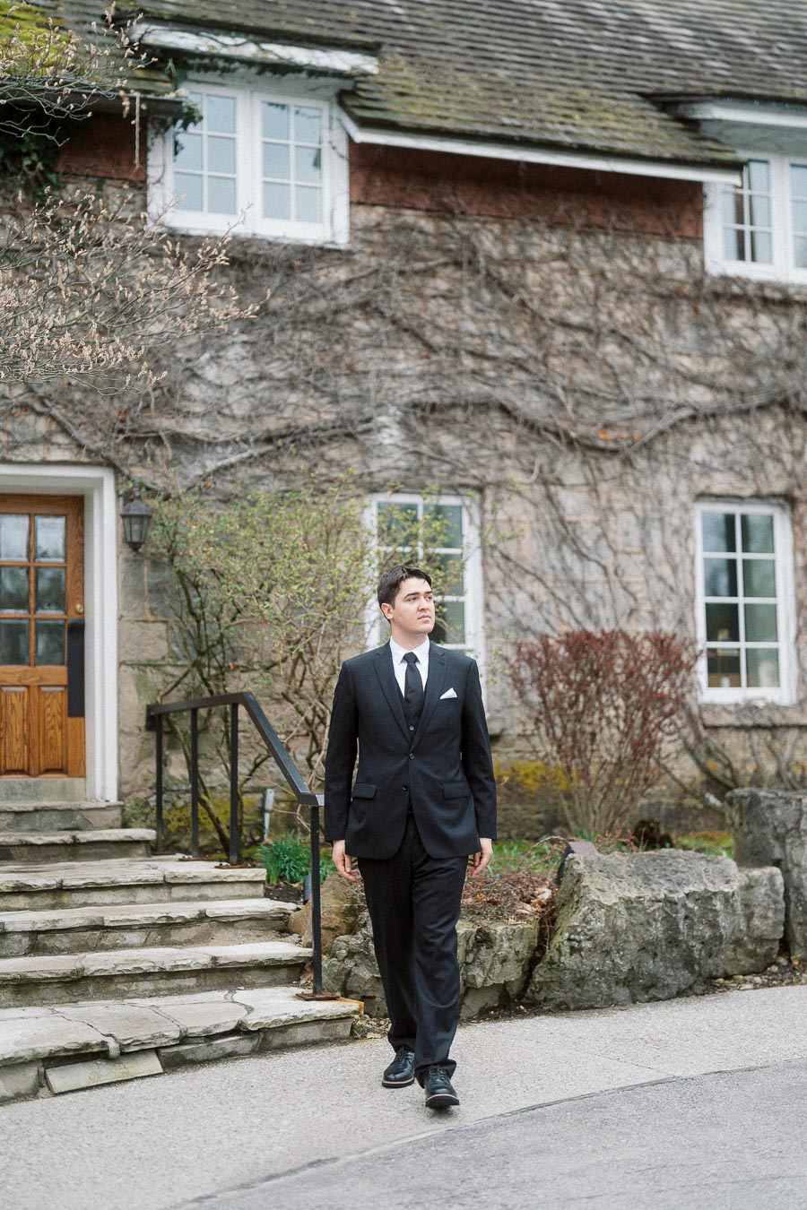 Man in a black suit walking outside a vine-covered stone building.