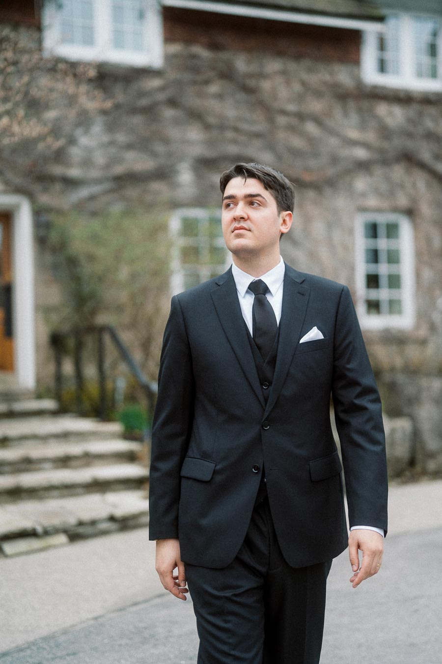 Man in a black suit standing outdoors near a stone building