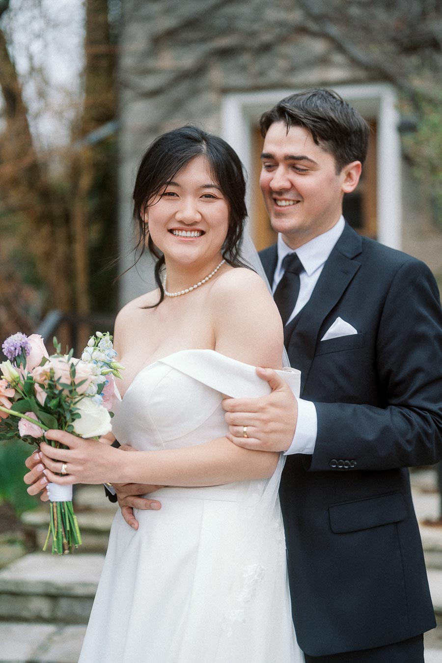 Couple in formal wedding attire smiling together outdoors, with the woman holding a bouquet.
