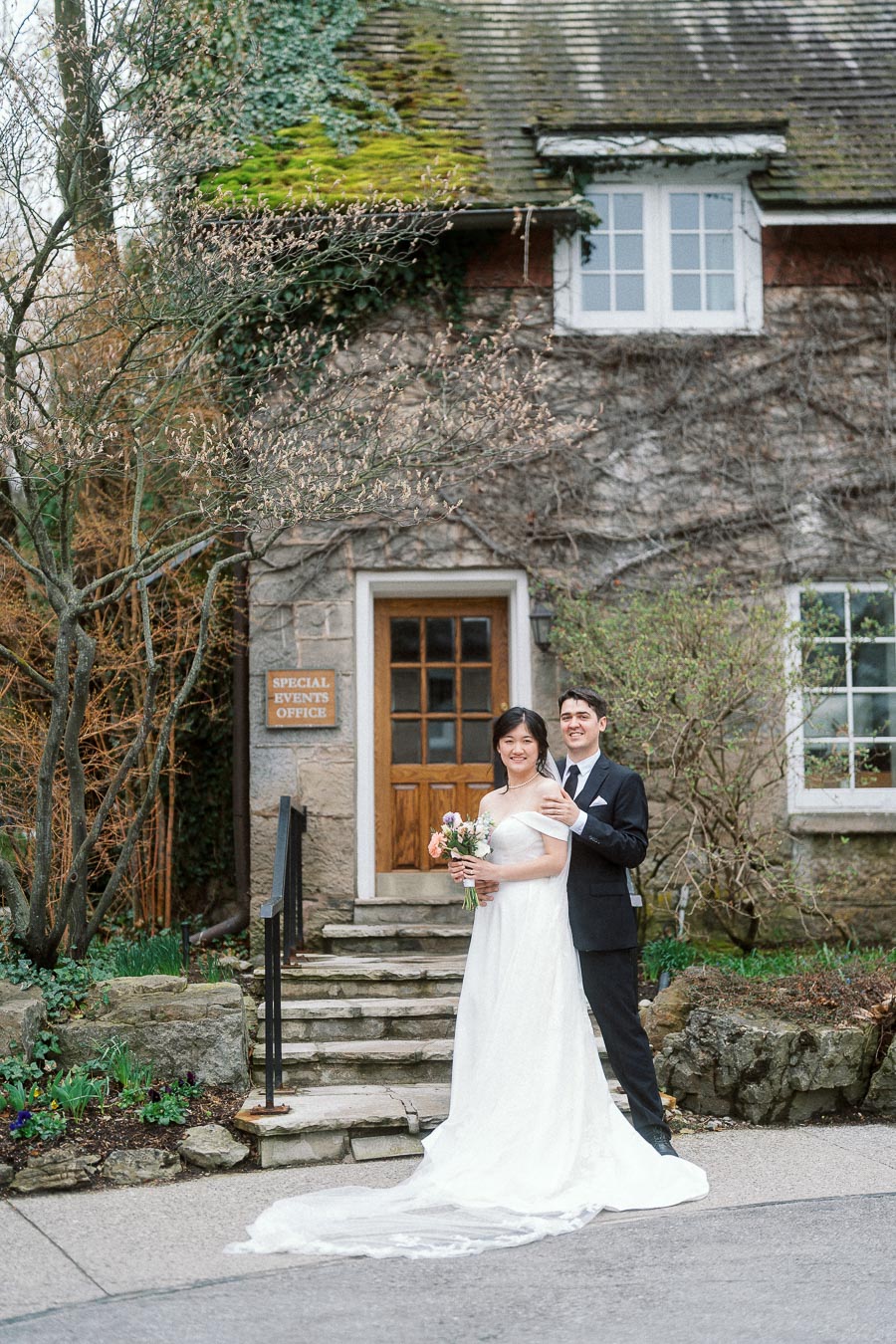 Couple in formal wedding attire posing outside a stone building with greenery