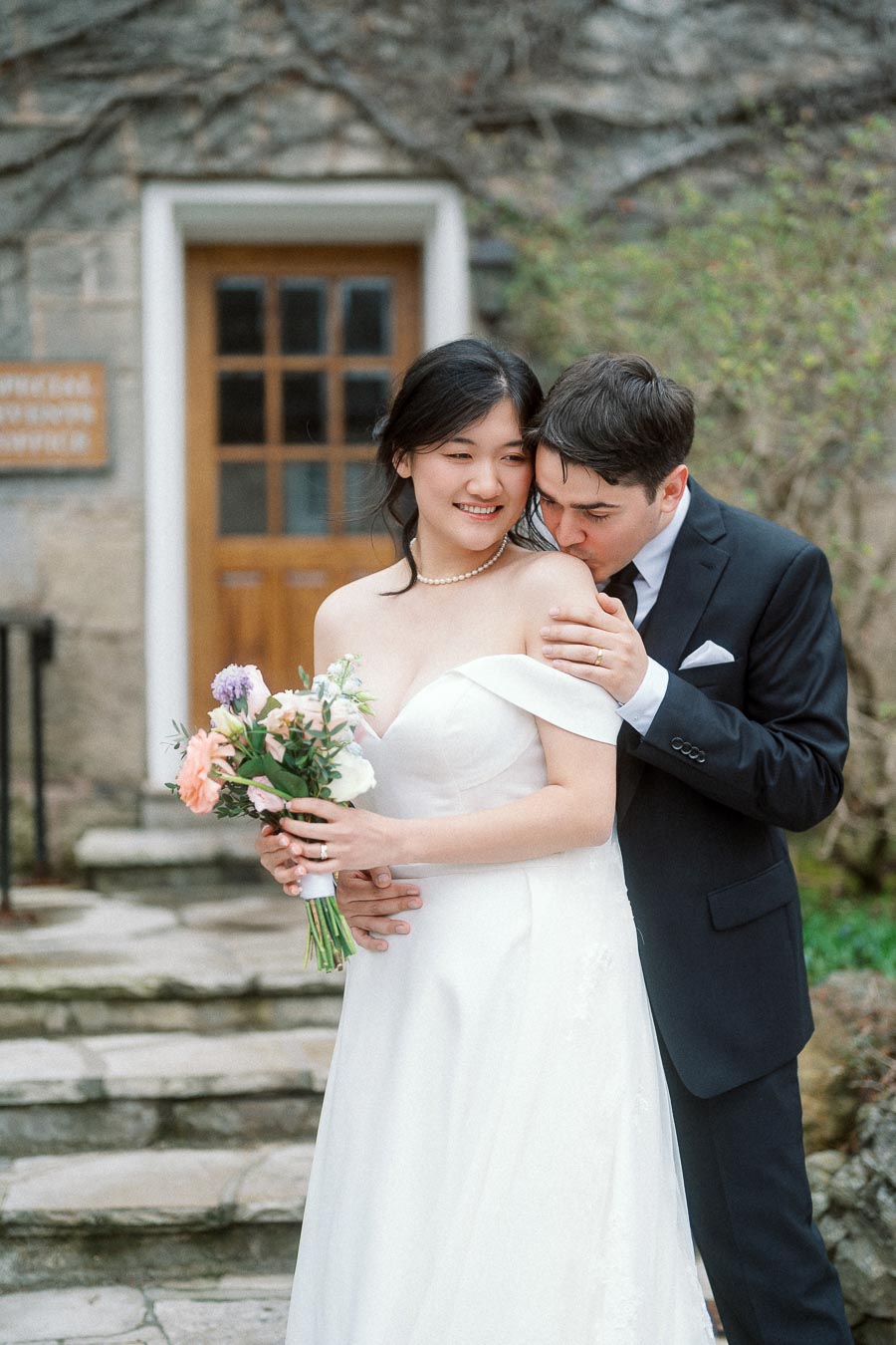 Groom kissing bride’s shoulder as she holds a bouquet on stone steps