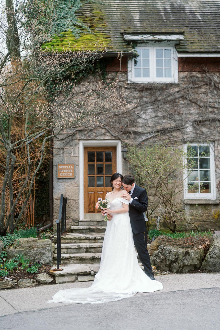 Couple in wedding attire posing outside a rustic stone building with greenery.