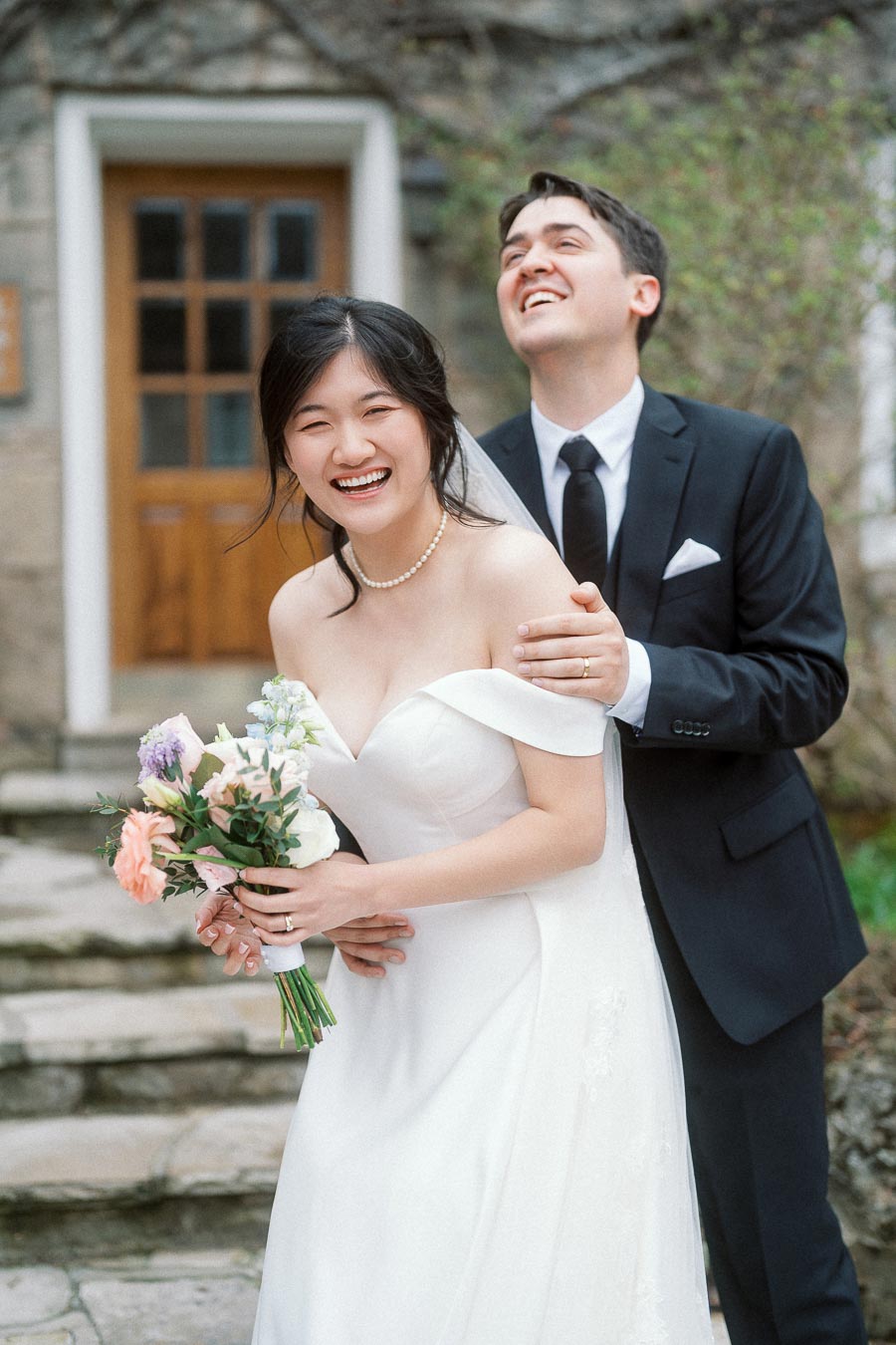 Laughing couple in formal attire with bouquet outside a stone building