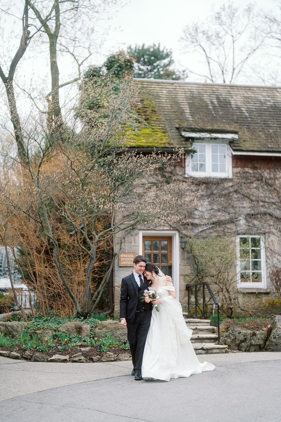 Couple in wedding attire walking arm in arm outside a rustic ivy-covered stone building