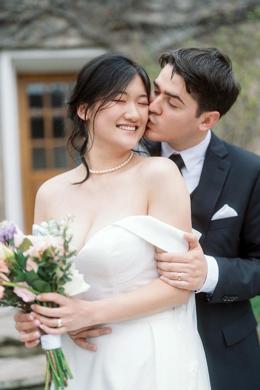 Groom kisses bride’s cheek as she smiles holding a bouquet outdoors