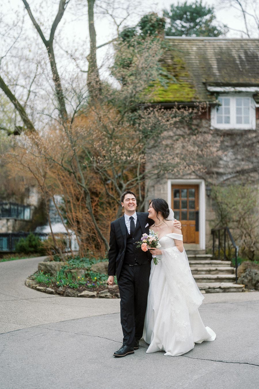 Couple in formal wedding attire walking arm in arm outside a vine-covered stone building