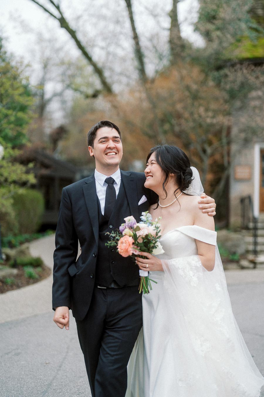 Couple in wedding attire walking arm in arm, smiling outdoors with a bouquet.