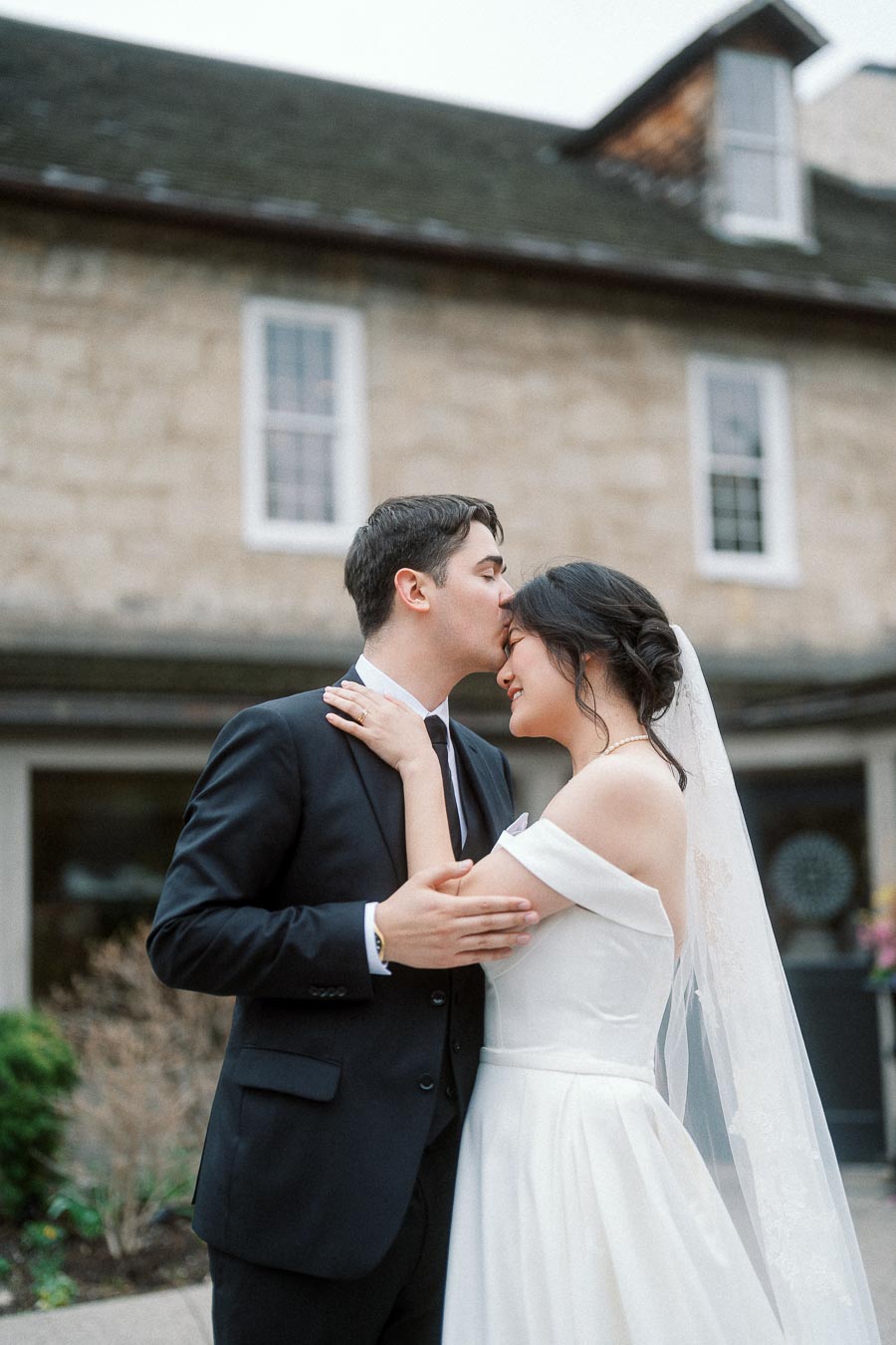 Groom kisses bride’s forehead outside a stone building.
