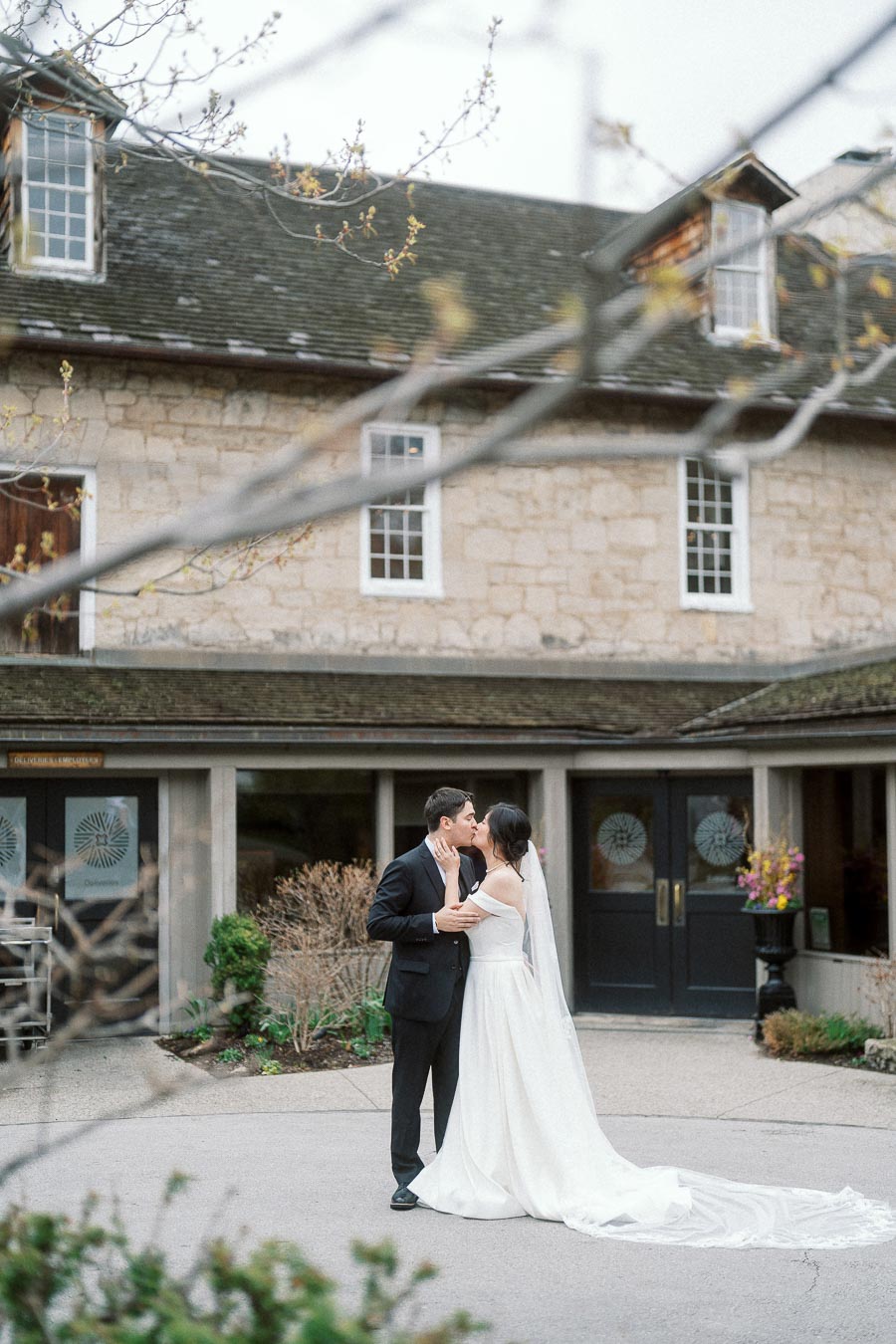 Couple in wedding attire kissing outside a stone building with tree branches in foreground