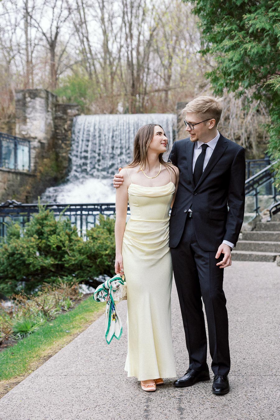 Couple in formal outfits walking arm in arm near a waterfall.