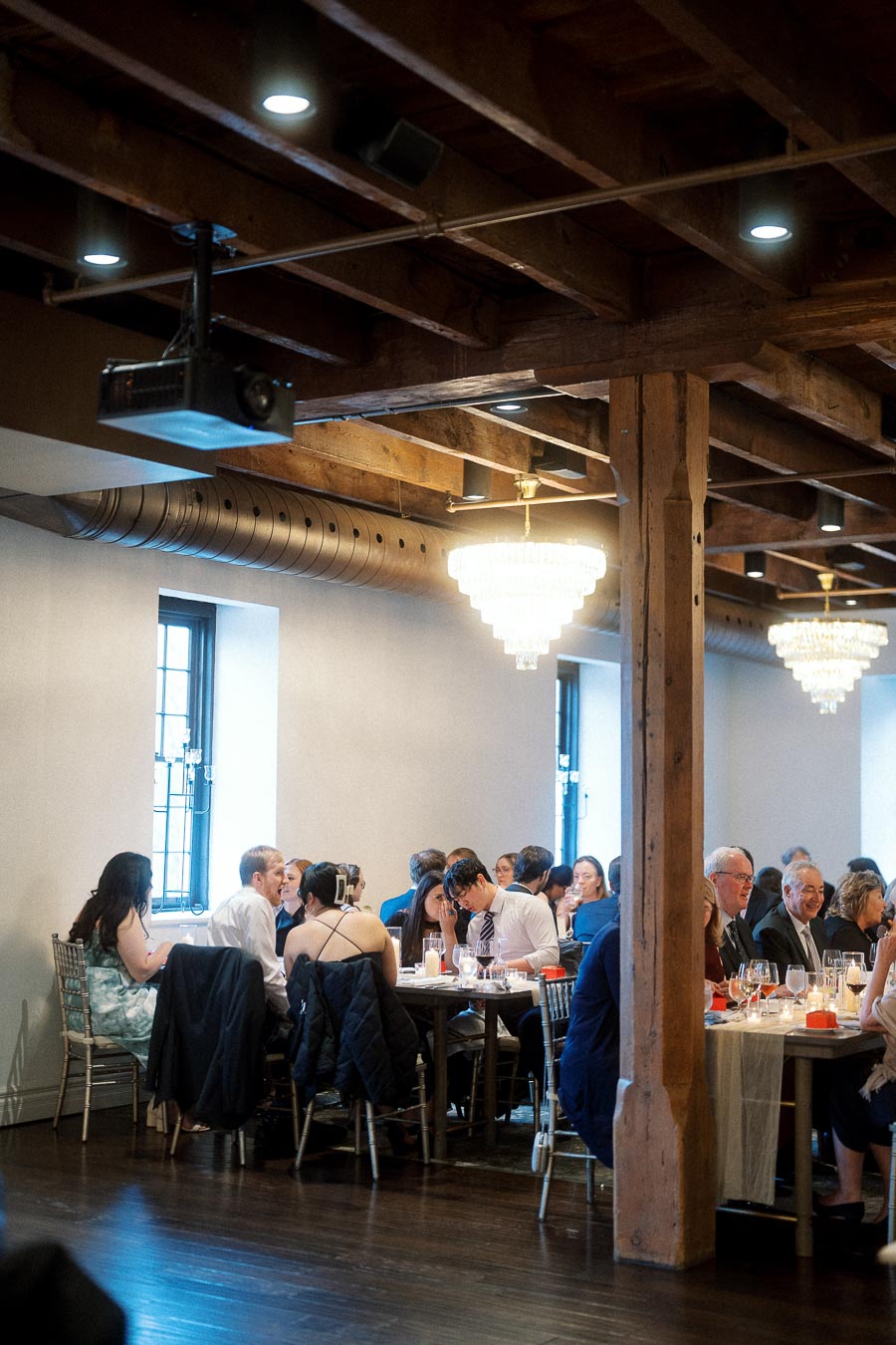 Guests seated at long tables in a rustic indoor event space with chandeliers