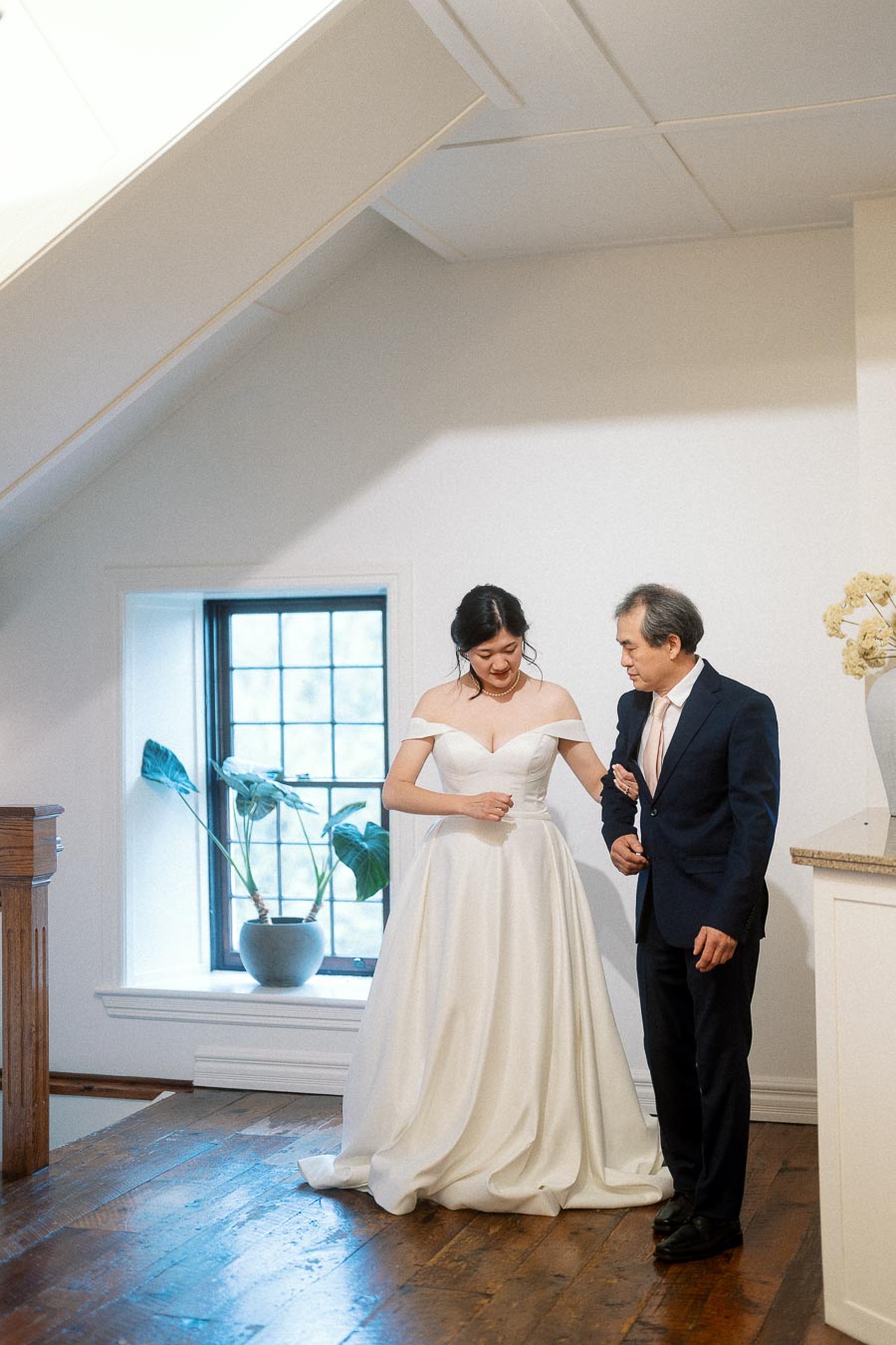 Bride in an off-the-shoulder gown walks arm-in-arm with an older man indoors