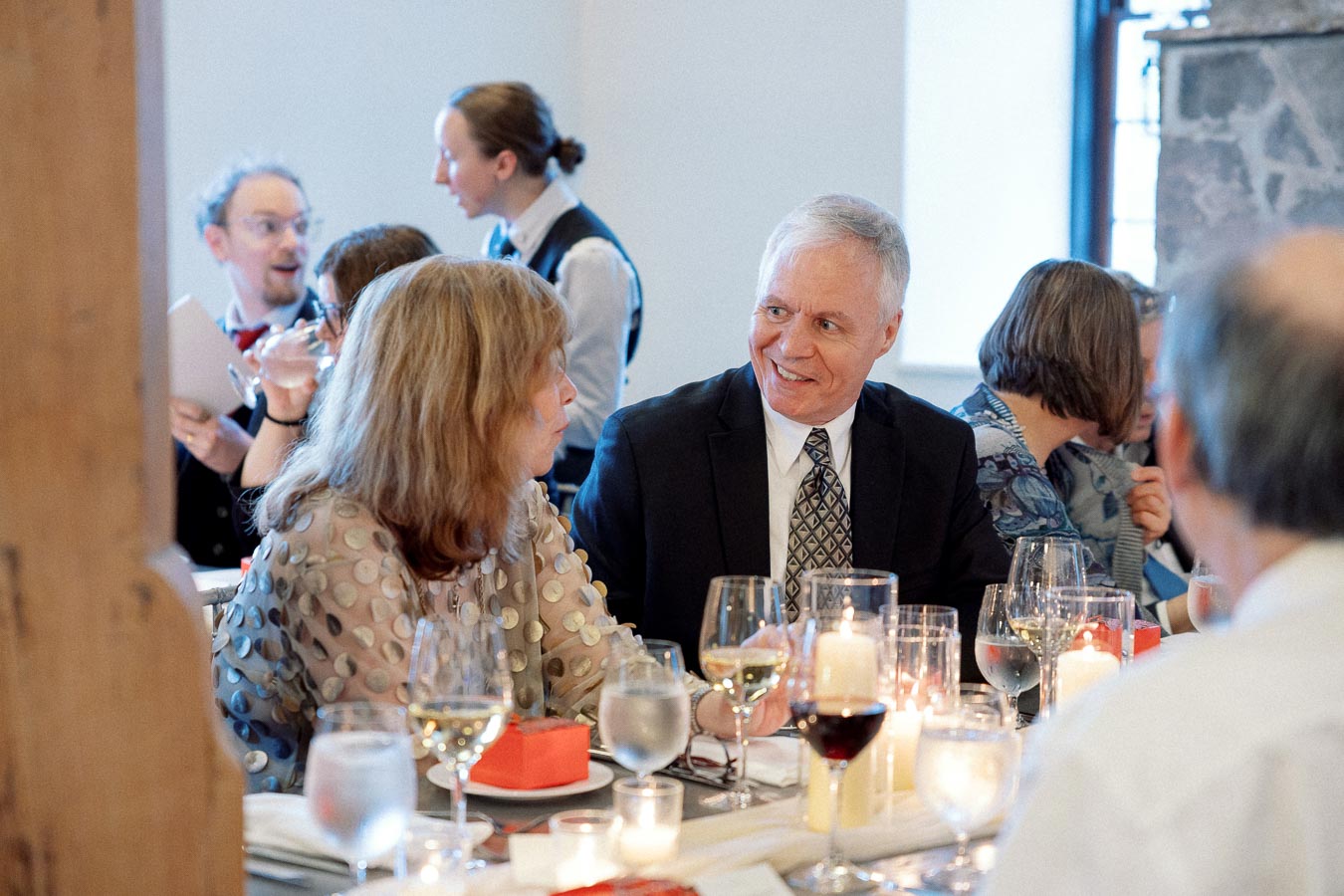 Guests converse at a candlelit dinner table with wine glasses and place settings.