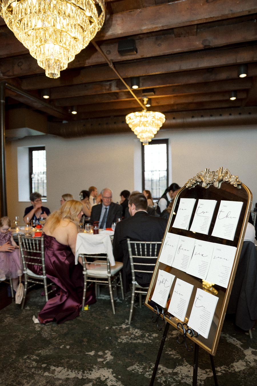 Reception seating chart displayed beside guests dining under chandeliers