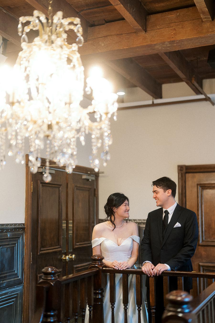 Couple in formal attire smiling on an indoor staircase beneath a chandelier