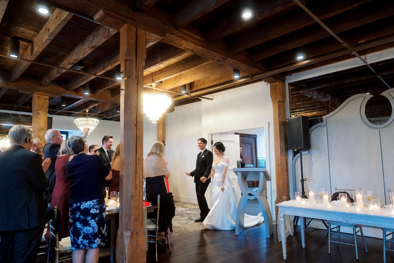 Couple in formal wedding attire walking into a candlelit reception as guests applaud