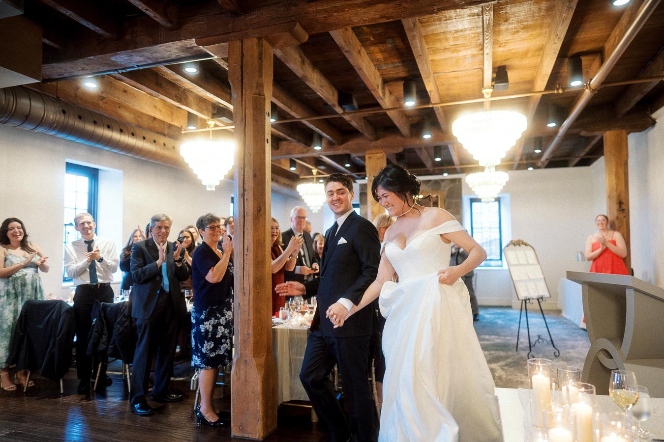 Bride and groom walk hand in hand into a reception as guests applaud.