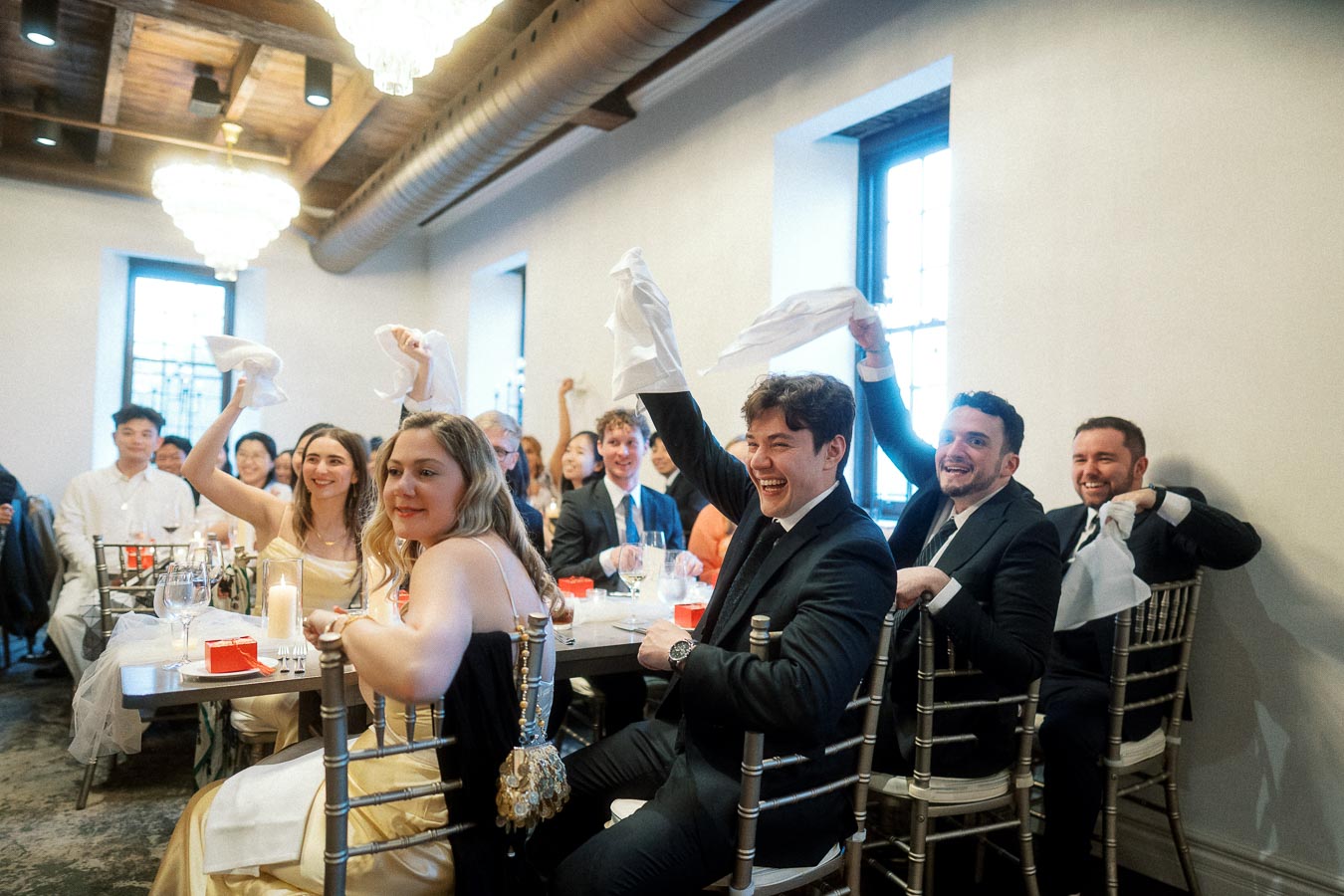 Guests seated at reception tables wave napkins and smile in a bright event room.