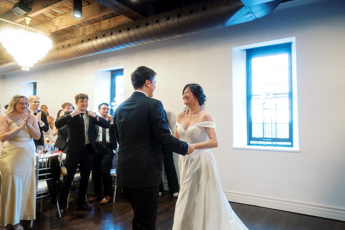 Bride and groom smiling during an indoor reception as guests clap nearby