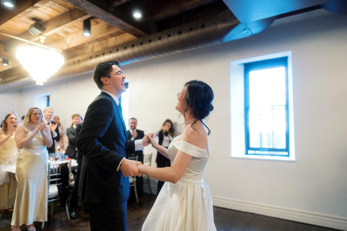 Couple laughing and holding hands while dancing indoors as guests applaud nearby