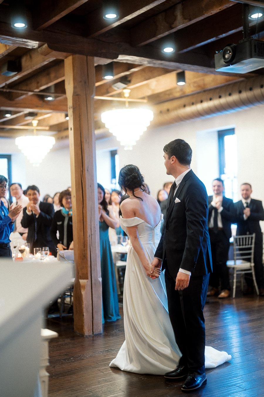 Bride and groom holding hands during a wedding reception as guests applaud.