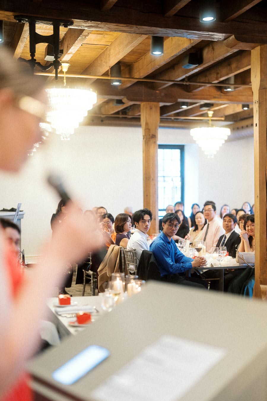 Guests seated at banquet tables listen to a speaker in a warmly lit event room.