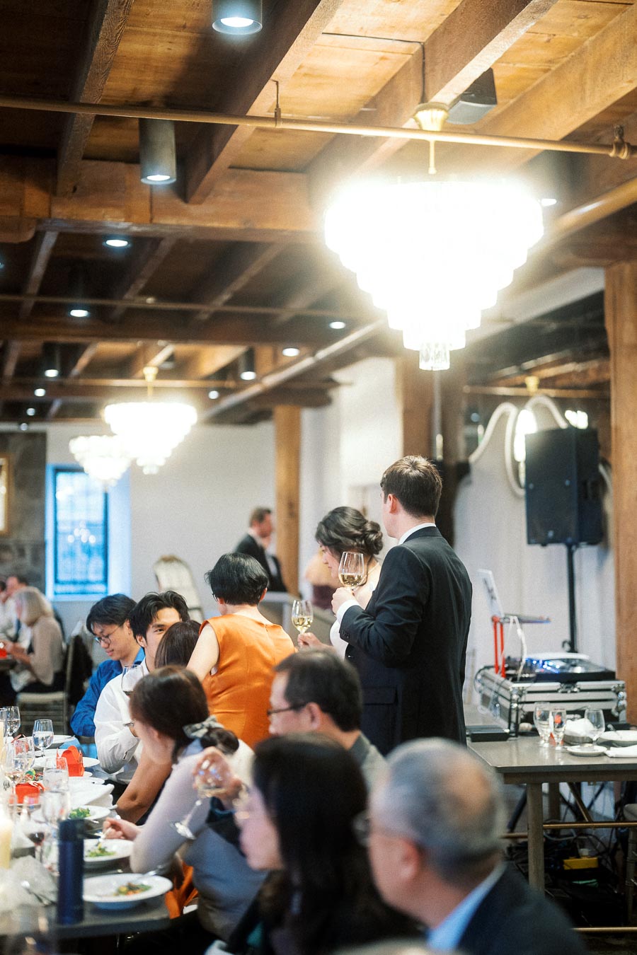 Guests seated at a formal indoor dinner beneath chandeliers and wooden ceiling beams