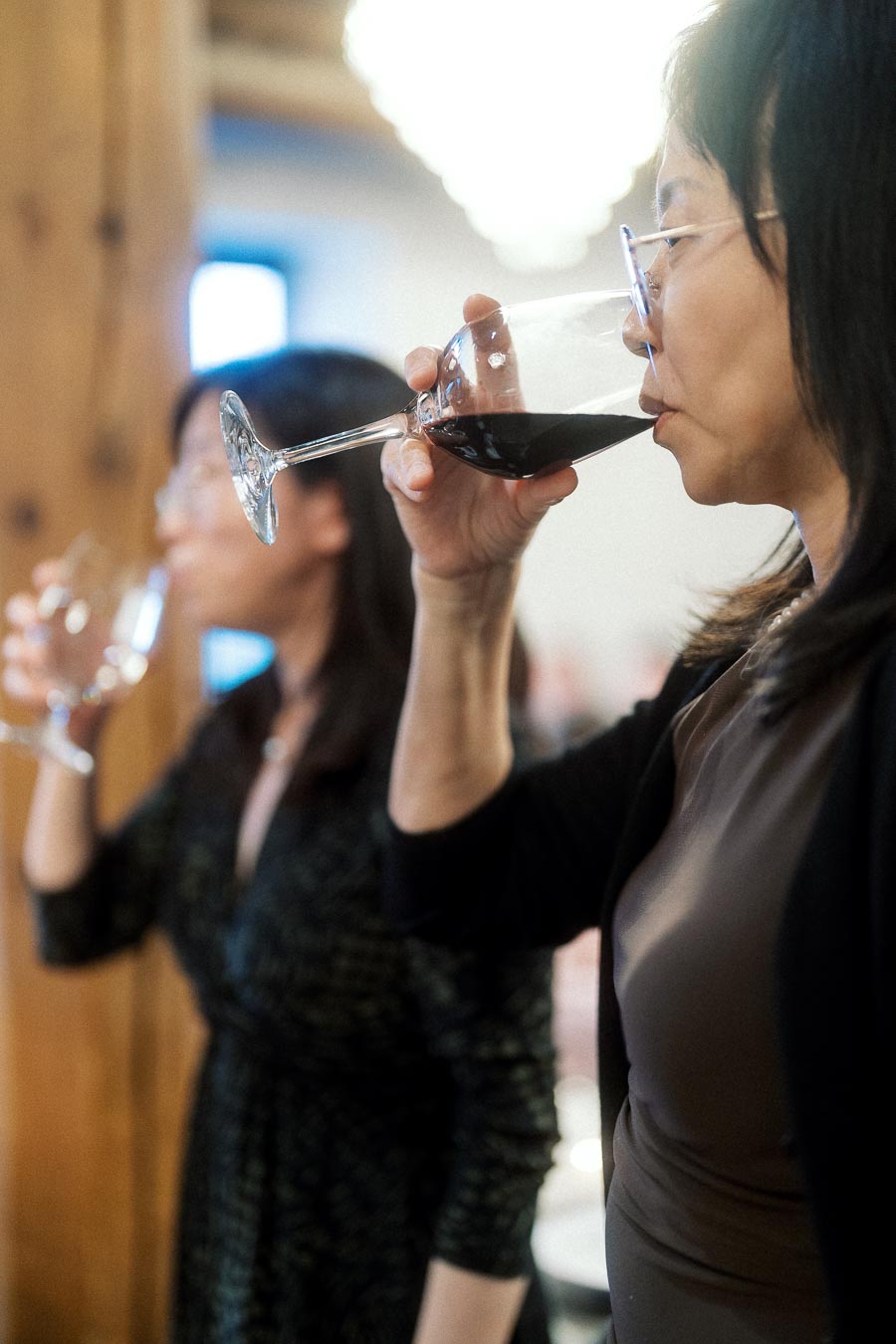 Two women tasting wine indoors, one in focus sipping from a glass