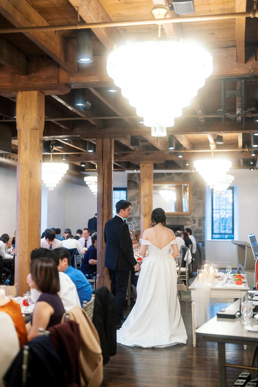 Couple in formal wedding attire walking through a warmly lit reception space