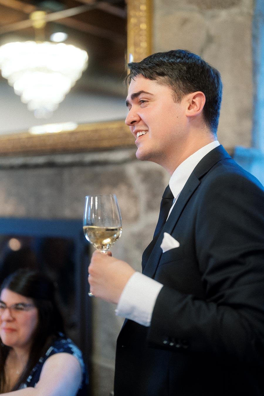 Man in a black suit smiling while holding a wine glass indoors