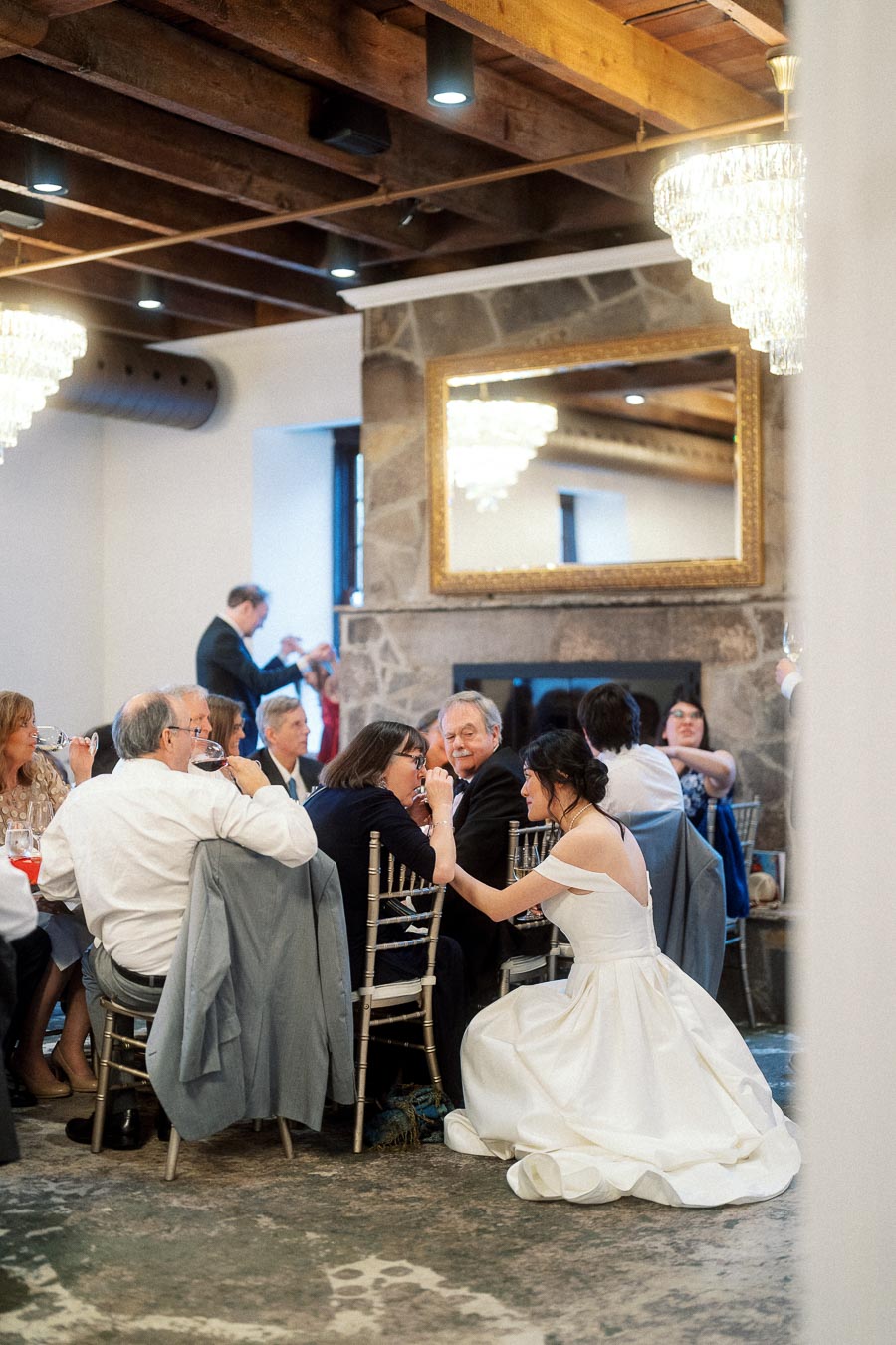 Bride kneeling beside seated guests during an indoor reception dinner.