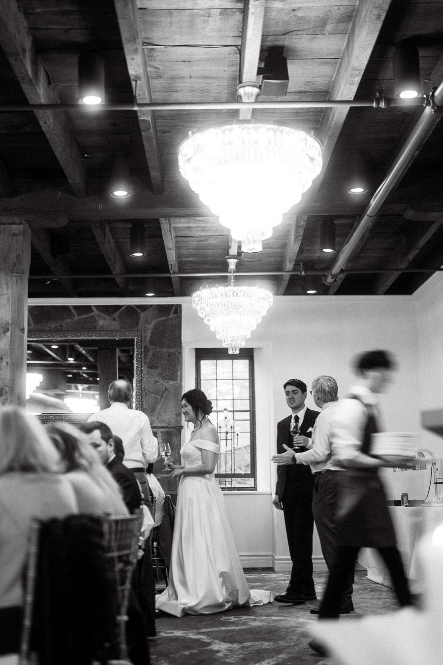 Couple in formal attire mingling with guests beneath chandeliers indoors
