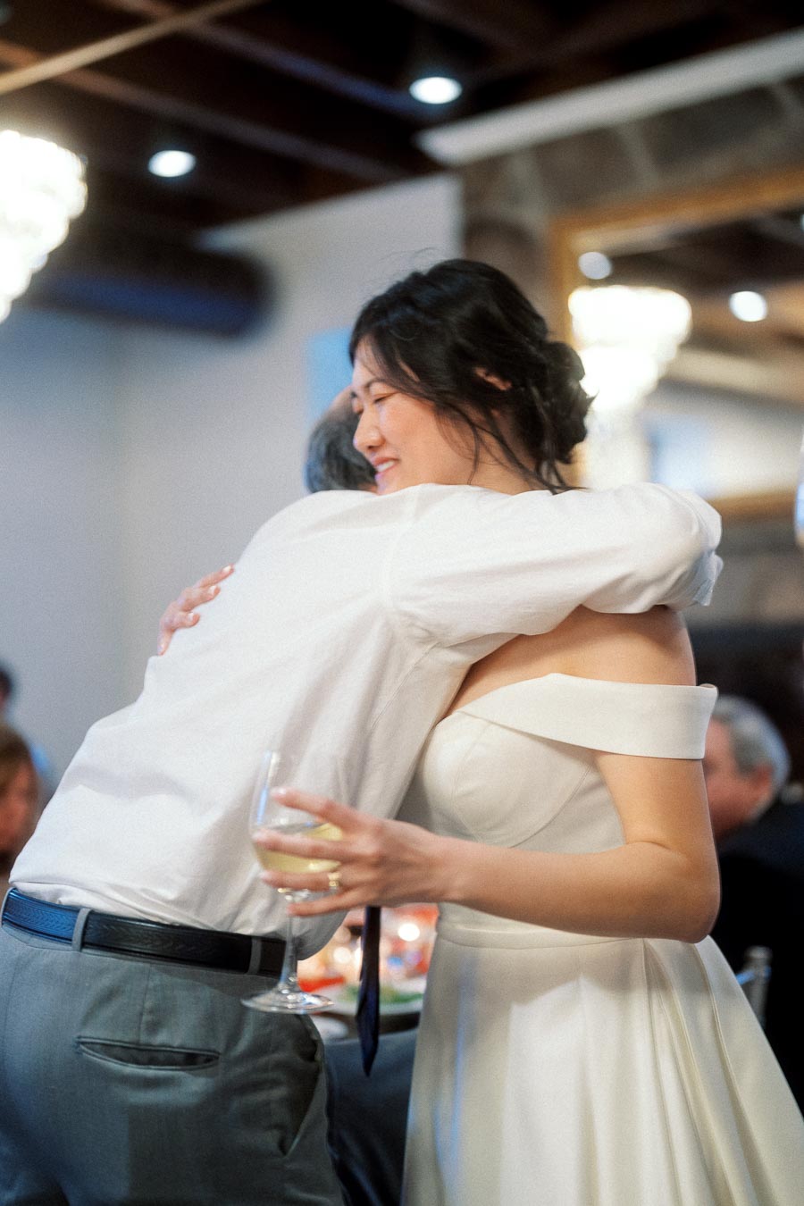 Woman in a white dress hugging a man while holding a wine glass indoors