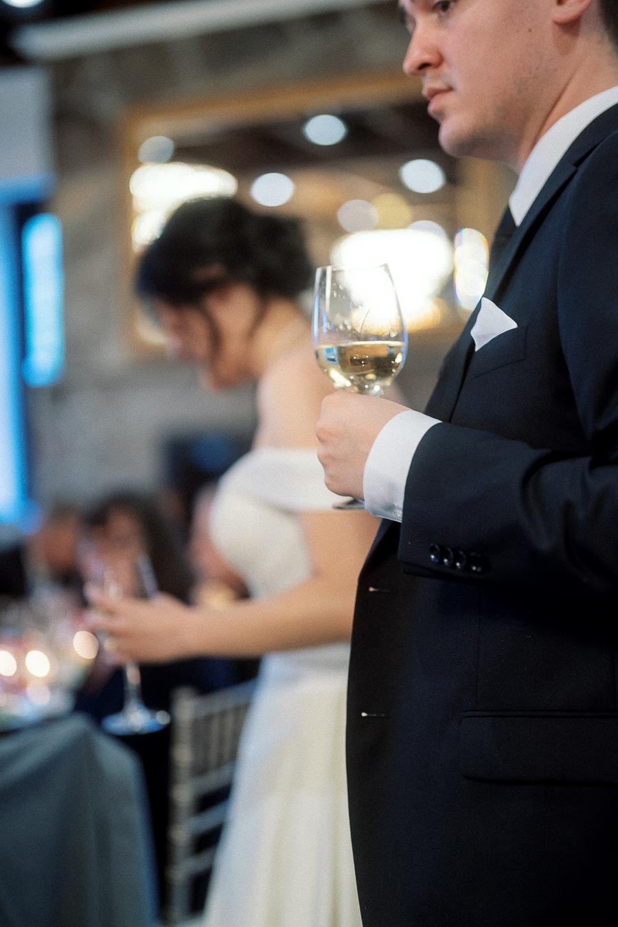 Man in a black suit holds a wine glass beside a woman in a white dress.